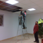 on aperçoit le maire qui regarde un employé de la mairie poser une plaque de plafond dans la salle des fêtes