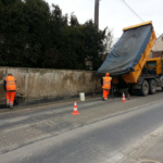 deux employés vêtus d'une combinaison de sécurité orange. l'un d'entre eux tasse le goudron avec un rouleau compresseur.